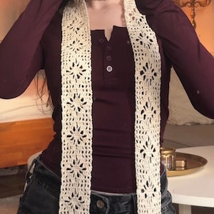 May include: Cream-colored, open-weave crochet scarf with a floral pattern. The scarf is long and rectangular, with a repeating design. The scarf is worn over a burgundy long-sleeved shirt and dark denim jeans.