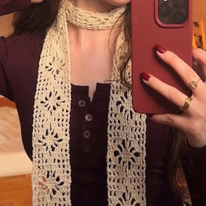 Puede incluir: Una bufanda de crochet color crema con un patrón floral calado. La bufanda está drapeada sobre una parte superior burdeos. El diseño de la bufanda presenta motivos florales repetidos, creando un accesorio aireado y decorativo.