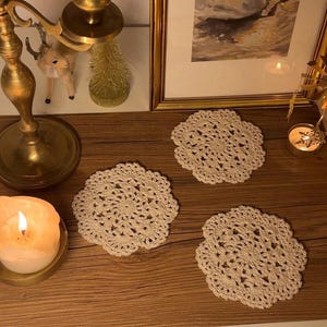 Könnte beinhalten: Vier cremefarbene, gehäkelte Untersetzer auf einer Holzoberfläche. Die Untersetzer haben ein kreisförmiges, durchbrochenes Design. Der Hintergrund umfasst eine brennende Kerze, einen goldenen Kerzenständer und ein gerahmtes Bild.