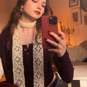 May include: A long, cream-colored crochet scarf featuring a repeating floral lace pattern. The scarf is worn wrapped around the neck with two long, vertical panels hanging down the front, showcasing intricate openwork stitches.