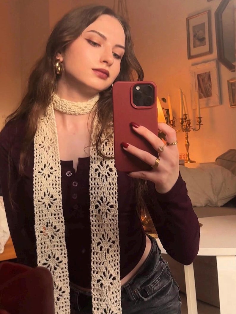 May include: A long, cream-colored crochet scarf featuring a repeating floral lace pattern. The scarf is worn wrapped around the neck with two long, vertical panels hanging down the front, showcasing intricate openwork stitches.