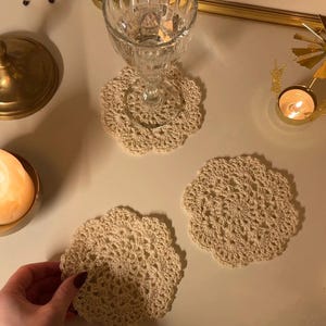 May include: Beige crocheted coasters with a floral design. One coaster supports a clear glass. The coasters are handmade and add a touch of elegance to any table setting. The coasters are approximately 5 inches in diameter.