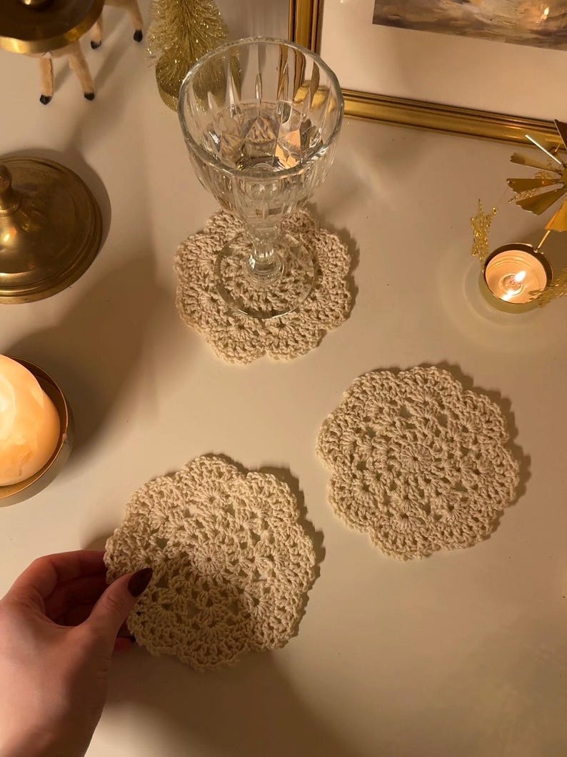 May include: Beige crocheted coasters with a floral design. One coaster supports a clear glass. The coasters are handmade and add a touch of elegance to any table setting. The coasters are approximately 5 inches in diameter.