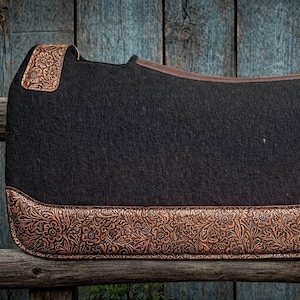May include: A black saddle pad with brown floral embossed leather accents. The pad has a rectangular shape with rounded corners and a brown leather trim. The embossed leather is on the corners and bottom edge.