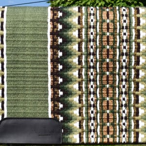 May include: A green and brown saddle pad with a geometric pattern. The pad features a central design of alternating brown, white, and green shapes, bordered by a solid green edge. A black leather piece is attached to the bottom.