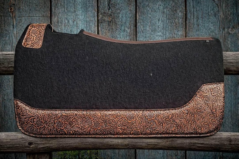 May include: A black saddle pad with brown floral embossed leather accents. The pad has a rectangular shape with rounded corners and a brown leather trim. The embossed leather is on the corners and bottom edge.