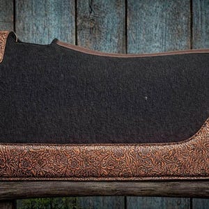 May include: A black saddle pad with brown floral embossed leather accents. The pad has a rectangular shape with rounded corners and a brown leather trim. The embossed leather is on the corners and bottom edge.