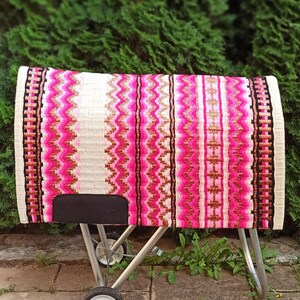 May include: A colorful horse saddle pad with a vibrant design. The pad features a white base with vertical stripes of pink, black, and gold. The edges are finished with a black and white border. The pad is displayed on a metal stand.
