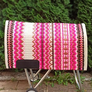 May include: A colorful horse saddle pad with a vibrant design. The pad features a white base with vertical stripes of pink, black, and gold. The edges are finished with a black and white border. The pad is displayed on a metal stand.