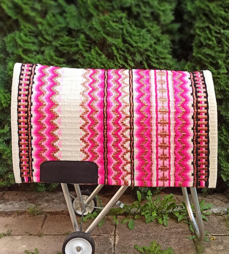 May include: A colorful horse saddle pad with a vibrant design. The pad features a white base with vertical stripes of pink, black, and gold. The edges are finished with a black and white border. The pad is displayed on a metal stand.