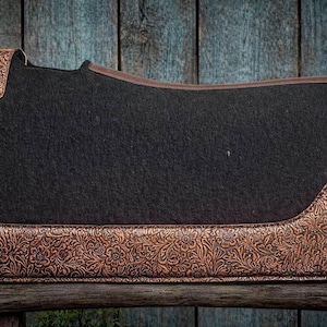 May include: A black saddle pad with brown floral embossed leather accents. The pad has a rectangular shape with rounded corners and a brown leather trim. The embossed leather is on the corners and bottom edge.
