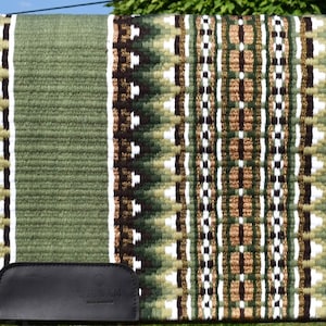 May include: A green and brown saddle pad with a geometric pattern. The pad features a central design of alternating brown, white, and green shapes, bordered by a solid green edge. A black leather piece is attached to the bottom.