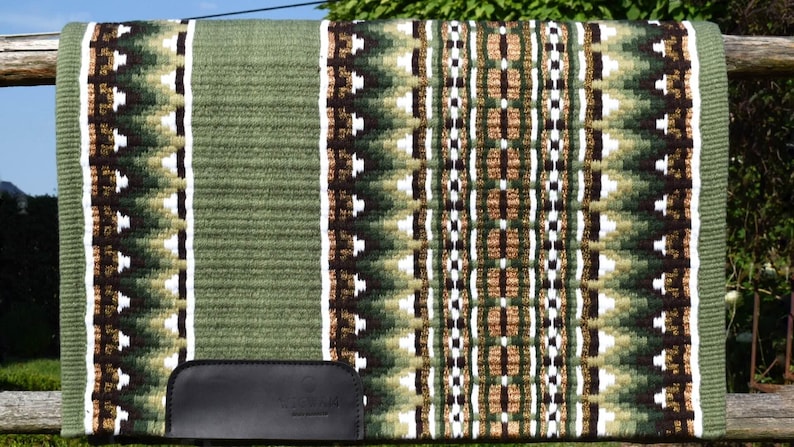 May include: A green and brown saddle pad with a geometric pattern. The pad features a central design of alternating brown, white, and green shapes, bordered by a solid green edge. A black leather piece is attached to the bottom.