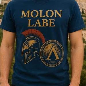 以下が含まれることがあります： 紺色のTシャツ。金色の文字で「MOLON LABE」と書かれています。デザインには、金色のスパルタンヘルメットと、金色のラムダシンボルが付いた盾が含まれています。背景には建物が見えます。
