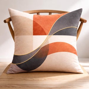 May include: A square throw cushion featuring a geometric pattern of colour-blocked sections in shades of beige, burnt orange, and charcoal grey. The design is accented by a thin, curved gold-coloured piping that sweeps across the surface.