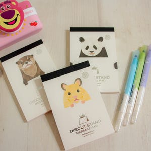 May include: A selection of Diecut Stand Message Pads with animal designs, such as a panda, otter, and hamster. A pink camera with a bear motif and a set of highlighters in various colours are also present.