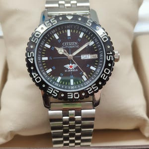 Op de afbeelding: Citizen automatisch horloge van roestvrij staal met een zwarte draaibare ring en een donkerbruine wijzerplaat. Het horloge heeft een datumweergave en een zilveren schakelband. Het horloge wordt getoond op een beige kussen.