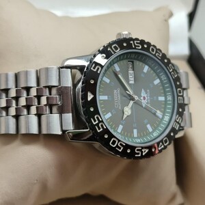 May include: A silver Citizen automatic watch with a black rotating bezel and a gray face. The watch has a stainless steel band and displays the day of the week. The watch is on a beige surface.