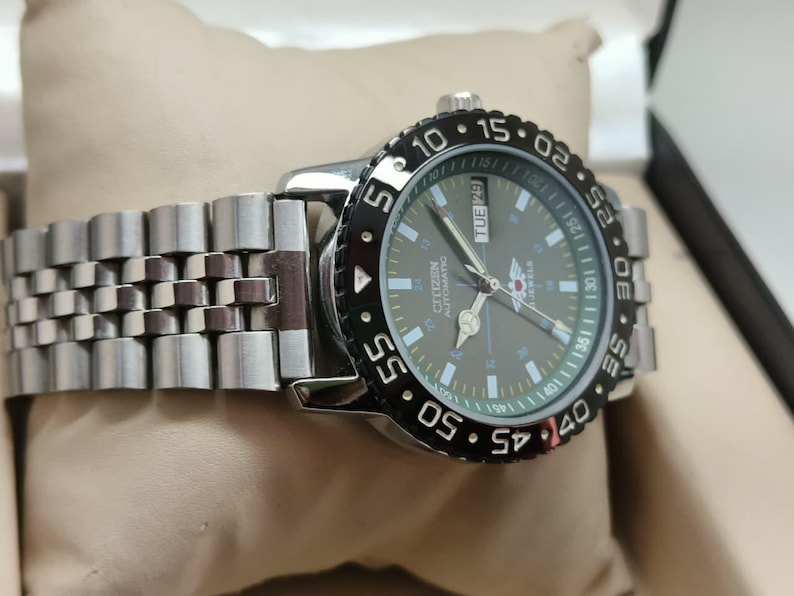 May include: A silver Citizen automatic watch with a black rotating bezel and a gray face. The watch has a stainless steel band and displays the day of the week. The watch is on a beige surface.