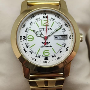May include: A gold-toned Citizen automatic wristwatch with a white face. The watch features a day and date display, green numerals, and a compass-style bezel. The watch band is also gold-toned.
