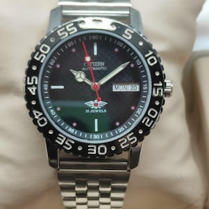 May include: A Citizen automatic wristwatch with a black face, silver hands, and a red second hand. The watch has a stainless steel strap and a black rotating bezel with white numerals. The day is displayed as "MON 20".