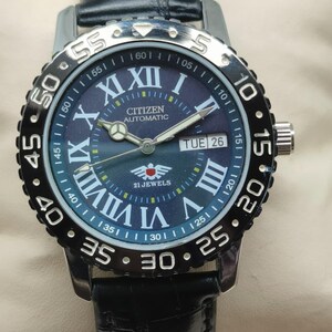 Può includere: Un orologio Citizen Automatic con cinturino in pelle nera. Il quadrante è blu scuro con numeri romani e lancette bianche. La lunetta è nera con segni bianchi. L'orologio mostra "TUE 26" e "21 JEWELS".