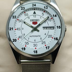 May include: Silver-toned Seiko automatic wristwatch with a white face. The watch features black numerals, red accents, and a day-date display. The watch has a mesh band and a stainless steel case.