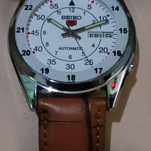 May include: A Seiko automatic wristwatch with a white face, black numerals, and luminous hands. The watch has a stainless steel case and a brown leather strap. The day is displayed at the 3 o'clock position.