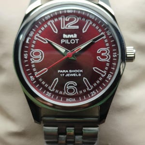 Op de afbeelding: Een polshorloge met een bordeauxrode wijzerplaat en een zilverkleurige kast en band. De wijzerplaat heeft witte cijfers, een seconden track en de tekst "humk PILOT PARA SHOCK 17 JEWELS INDIA". Het horloge heeft een zilverkleurige kroon.