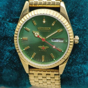 May include: A gold-tone Citizen automatic wristwatch featuring a round green dial with gold-tone hour markers, a day-date window at the three o'clock position, a red second hand, and a gold-tone metal link bracelet with a textured finish.