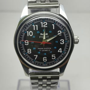 May include: A silver wristwatch with a black face. The watch features white numerals, a red second hand, and the text "PILOT" and "PARA SHOCK 17 JEWELS". The watch has a stainless steel band.