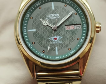 May include: Gold-toned Citizen automatic wristwatch with a green dial and gold hands. The watch face displays the day and date, with a patterned center and a gold bezel. The watch band is also gold-toned.