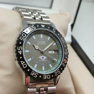 May include: A silver Citizen automatic watch with a black and silver bezel. The watch face is gray with white and green markings, and the day is displayed as "TUE 29". The watch has a silver link band.