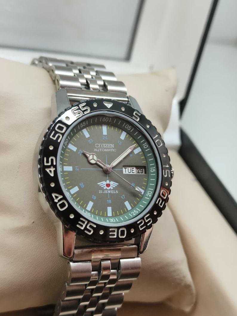 May include: A silver Citizen automatic watch with a black and silver bezel. The watch face is gray with white and green markings, and the day is displayed as "TUE 29". The watch has a silver link band.