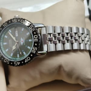 May include: A silver wristwatch with a black rotating bezel and a dark green face. The watch has a silver band and is displayed on a beige pillow. The watch face features white and silver markings.