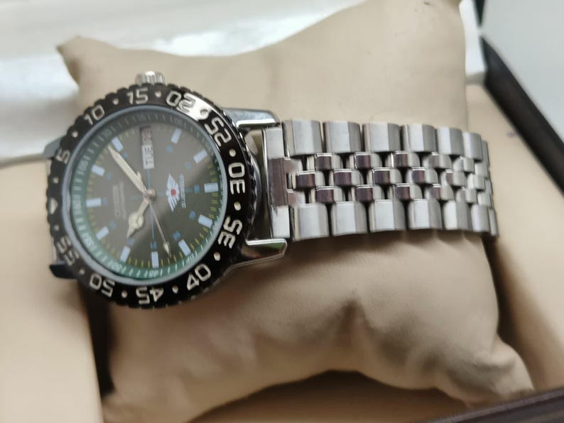 May include: A silver wristwatch with a black rotating bezel and a dark green face. The watch has a silver band and is displayed on a beige pillow. The watch face features white and silver markings.