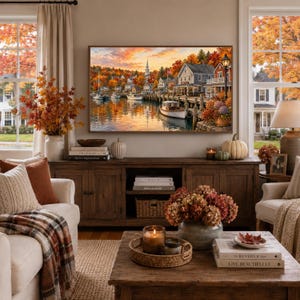 May include: A framed landscape painting of a harbor town during autumn, featuring boats docked at a wooden pier, colorful fall foliage, and a church steeple. The artwork is displayed above a wooden media console in a living room setting.