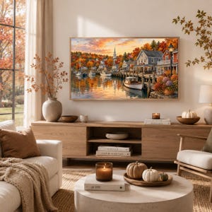 May include: A framed landscape painting depicting a coastal village at sunset with boats docked at a wooden pier, surrounded by autumn foliage in shades of orange and red. The scene includes a church steeple, houses, and a calm harbor reflecting the sky.