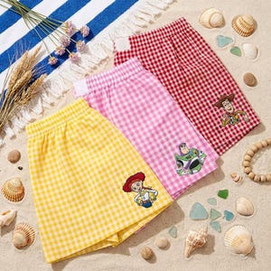 May include: Three pairs of elastic-waist shorts in yellow, pink, and red gingham patterns. Each pair features a small embroidered character from the Toy Story franchise: Jessie on the yellow shorts, Buzz Lightyear on the pink, and Woody on the red.