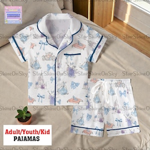 May include: White pajama set with a blue trim, featuring a pattern of castles, bows, and princess dresses. The set includes a short-sleeved top with a chest pocket and shorts with a drawstring. Text on the image reads: Adult/Youth/Kid Pajamas.