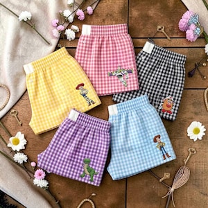May include: Five pairs of gingham shorts with elastic waistbands in yellow, pink, black, purple, and light blue. Each pair features a small embroidered character from the Toy Story franchise, including Woody, Buzz Lightyear, Mr. Potato Head, and Rex.