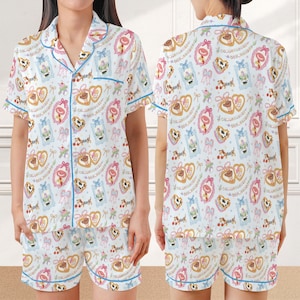 May include: A short-sleeved button-down pajama shirt and matching shorts set in white, featuring a repeating pattern of Toy Story characters, pink heart-shaped frames, blue bows, and cherries. The set includes blue piping along the edges.