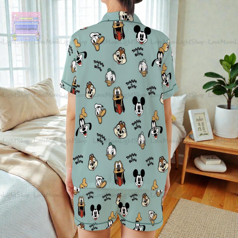 a person wearing a light blue pajama top with a pattern of cartoon characters, including Mickey Mouse, Goofy, Donald Duck, and other popular Disney characters. The person is standing in a bedroom with a bed, a nightstand, and a plant visible in the background.