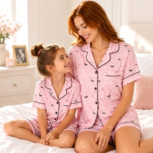 May include: Matching pink short-sleeve pajama sets featuring a button-down shirt with a notched collar and shorts. The fabric has a repeating pattern of small black bats and white ghosts. Each garment is finished with black piping along the edges.