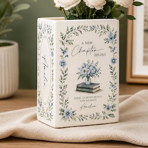 May include: A rectangular ceramic vase with a cream finish, decorated with blue floral vines and a central illustration of a bouquet on books. The text reads A NEW CHAPTER BEGINS, EVERY CHAPTER BRINGS NEW BEGINNGS, and the name Amelia appears twice.
