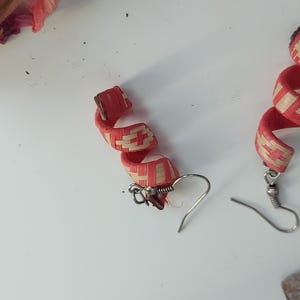 Könnte beinhalten: Spiralohrringe mit einem gewebten Muster in Rot und Beige. Die Ohrringe haben einen Hakenverschluss und werden auf einer weißen Oberfläche präsentiert. Das Design erinnert an ein gewebtes Band.