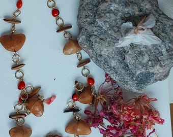 May include: A handmade necklace featuring brown, heart-shaped pendants, small red beads, and wooden accents. The necklace is connected by gold-colored chain links. The image also includes a gray rock and dried flowers.
