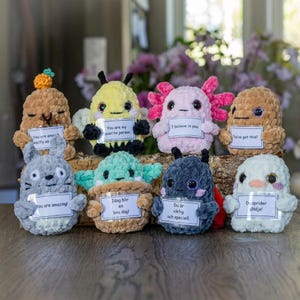 Peut inclure: Un ensemble de huit petites peluches crochetées de formes et de couleurs variées, incluant un axolotl rose, une créature grise et un personnage vert dans un pot. Chaque peluche tient un petit panneau blanc avec du texte imprimé en anglais ou suédois.