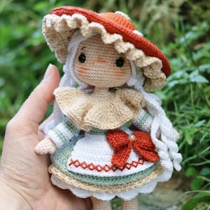 Puede incluir: Muñeca de ganchillo hecha a mano con un sombrero en forma de hongo. La muñeca tiene pelo blanco, un cuello beige y un vestido verde y blanco con un lazo rojo. La muñeca mide aproximadamente 15 cm de altura.