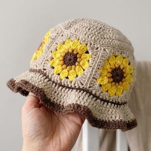 May include: A crocheted bucket hat in beige with three yellow and brown sunflower appliqués. The brim and edges are trimmed with a dark brown color. The hat is handmade and has a textured appearance.